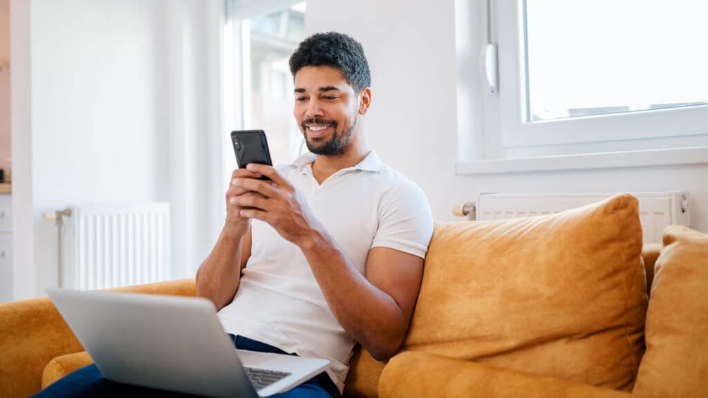 Smiling business owner using smartphone and laptop at home to manage calls with PATLive’s spam call blocking for business.